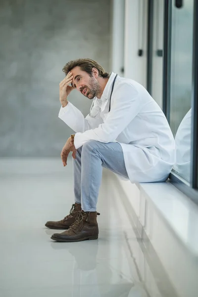 コヴィド19のパンデミックの間 空の病院の廊下の窓の近くに座っている間 ストレスを感じている成熟した医者のショット — ストック写真
