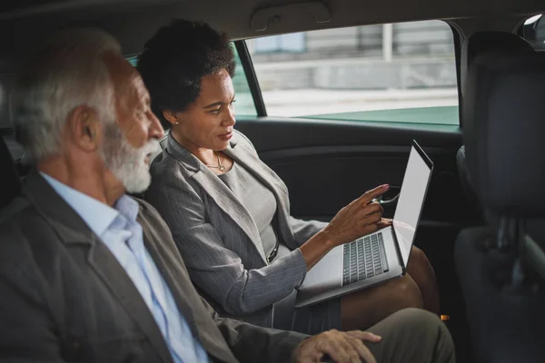2人の成功した多民族の人々のショットノートパソコンを使用して 彼らのビジネス通勤中に車の後部座席に座って議論を持っている — ストック写真