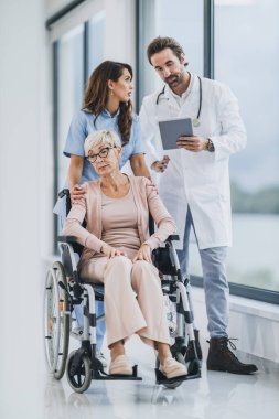 Başarılı bir doktor ve genç hemşire tekerlekli sandalyedeki yaşlı bir kadınla konuşuyor ve ilgileniyor..