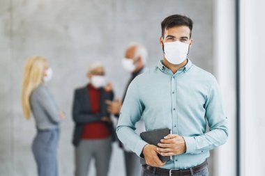 Korumalı maskeli başarılı genç bir işadamı dijital tablet tutuyor ve Corona virüsü salgını sırasında ofiste duruyor. Kameraya bakıyorum..