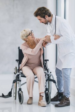 Genç bir doktorun, hastane koridorunda tekerlekli sandalyeye mahkum yaşlı bir kadına bakması..