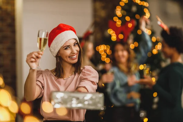 Happy young woman bringing presents and toasting with champagne as she celebrate Christmas with her friends at home party.