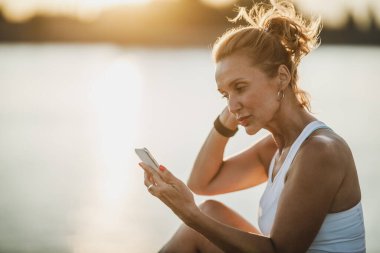 Nehrin kenarında oturan orta yaşlı bir sporcu gün batımında antrenmandan sonra dinlenirken akıllı telefonuyla sörf yapıyor.. 