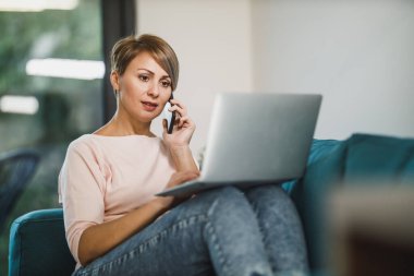 Orta yaşlı bayan girişimci oturma odasında otururken cep telefonuyla konuşuyor ve bir dizüstü bilgisayar üzerinde çalışıyor..