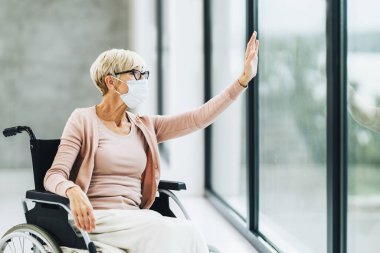 Engelli, koruyucu maskeli, tekerlekli sandalyede oturan ve Covid-19 salgını sırasında pencereden sallanan yaşlı bir kadın..