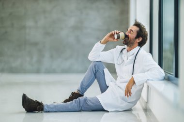 Gülümseyen bir doktor pencerenin yanında oturuyor ve boş bir hastane koridorunda kahve molası veriyor..