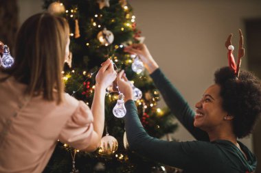 Çok ırklı güzel bayan arkadaşlar tatilin tadını çıkarıyorlar, Noel ağacı süslüyorlar, Noel ışıklarını düzenliyorlar ve eğleniyorlar..