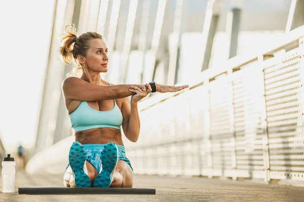 Middle age sports woman warming up and stretching her arms on a bridge.