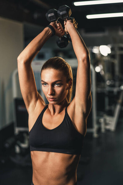 Молодая мускулистая женщина тренируется с гантелями для мышц трицепса в тренажерном зале.
