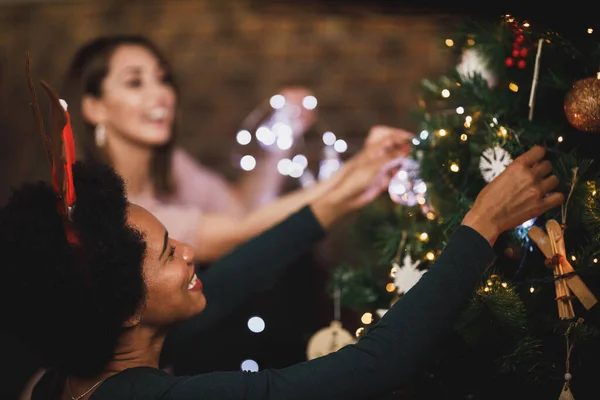 幸せなアフリカ系アメリカ人女性は家で休日の時間を楽しんで 彼女のクリスマスツリーを飾り 友人とクリスマスライトを配置します — ストック写真