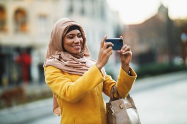 Siyahi Müslüman kadın tesettür takıyor ve akıllı telefonuyla şehir ortamında fotoğraf çekiyor..