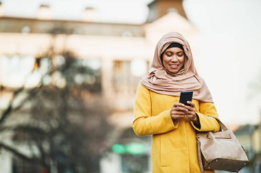 Siyahi Müslüman kadın tesettür takıyor ve şehir ortamında yürürken akıllı telefonuna mesajlar bırakıyor..