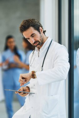 Dijital tablet kullanan yorgun bir doktor hastane koridorunda hızlı bir mola verirken saate bakıyor..