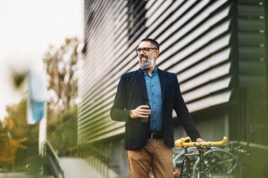 Başarılı bir orta yaşlı işadamı, yanında bir fincan kahveyle ve bisikletle, ofisin önünde yürüyor..