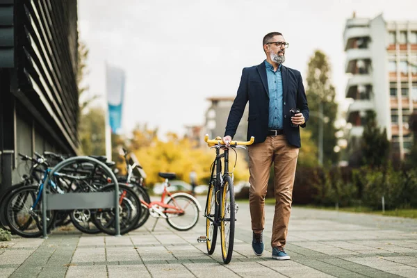 彼の横に自転車で行くためにコーヒーを飲みながら成功した中年のビジネスマン オフィス街の前を歩く — ストック写真