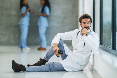 A worried doctor sitting on the floor and pensive looking out the window while having a quick coffee break in an empty hospital hallway.