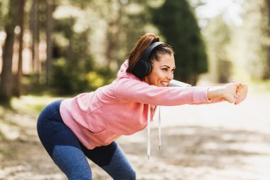 Genç, güzel, gülümseyen bayan koşucu doğada koşmadan önce ısınırken müzik dinliyor..
