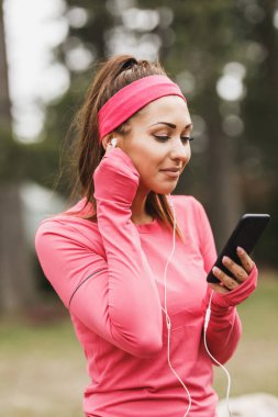 Genç, güzel, gülümseyen bayan koşucu müzik dinliyor ve doğada koştuktan sonra mola veriyor..