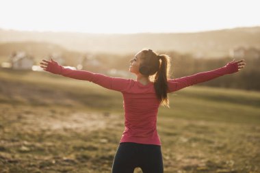 Genç, güzel, gülümseyen bayan koşucu doğada koştuktan sonra esneme hareketleri yaparken müzik dinliyor..