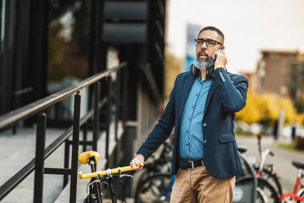 中年のビジネスマンが自転車で仕事に行き オフィス街の前のスマートフォンで話す — ストック写真