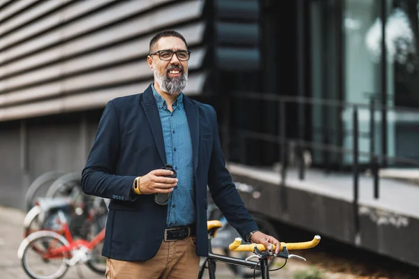 彼の横に自転車で行くためにコーヒーを飲みながら成功した中年のビジネスマン オフィス街の前を歩く — ストック写真