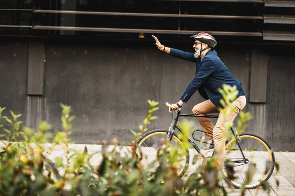 成功した中年のビジネスマンは誰かに手を振って自転車に乗って街を歩く — ストック写真