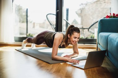 Young woman using laptop while doing plank exercises at home in the morning.