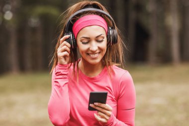 Genç, güzel, gülümseyen bayan koşucu müzik dinliyor ve doğada koşuya hazırlanıyor..