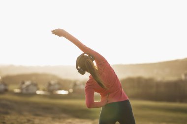 Genç, güzel, gülümseyen bayan koşucu doğada koştuktan sonra esneme hareketleri yaparken müzik dinliyor..
