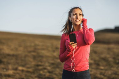 Genç, güzel, gülümseyen bayan koşucu müzik dinliyor ve doğada koştuktan sonra mola veriyor..