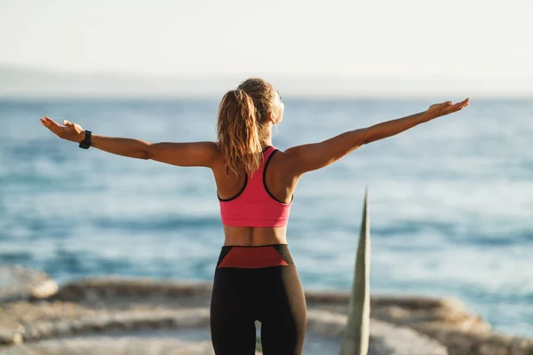 Happy young woman breathing deep fresh air and raising arms during stretching workout near the sea beach.