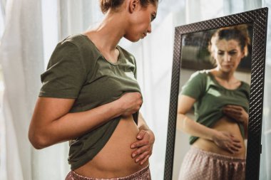 Frown young woman standing in front of a mirror and holding hands on her bloating stomach. clipart