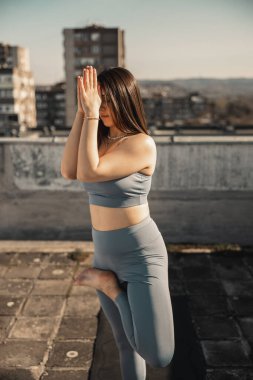 Terasta yoga yapan rahat genç bir kadın. Arka planda şehir binaları.