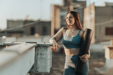 Relaxed woman is holding exercise mat and preparing to practicing yoga on a rooftop terrace.