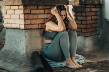 Lonely young woman in depression sit on a rooftop terrace lost in bad pessimistic thoughts after yoga training on a rooftop terrace.