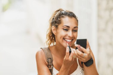 Portrait of a young smiling woman using hand mirror and applying lipstick while enjoying a summer vacation.