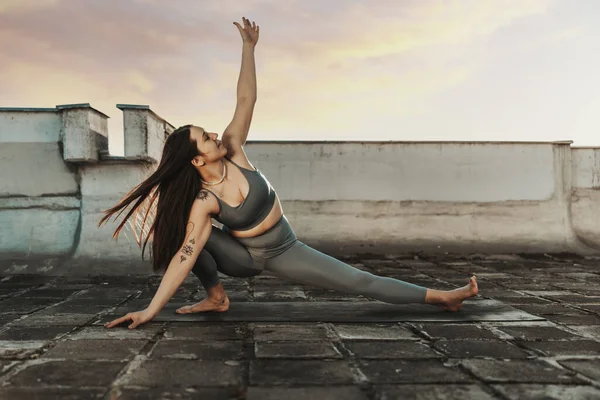 Relaxed woman practicing yoga stretching exercise on a rooftop terrace at sunset.
