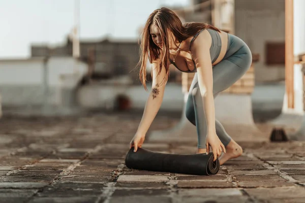 Relaxed woman is rolling up exercise mat and preparing to practicing yoga on a rooftop terrace.