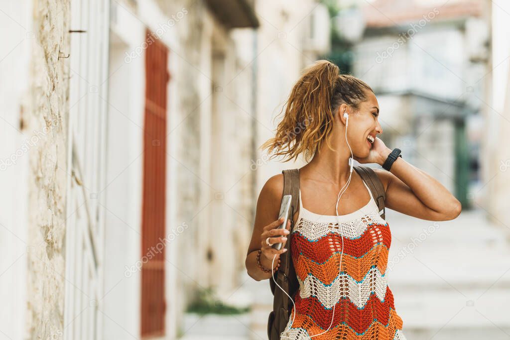 A young smiling woman enjoying music while exploring a Mediterranean city on a summer vacation.