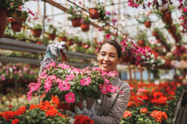 Smiling young woman working in a flower garden, holding flower pots and enjoying in beautiful and colorful flowers.