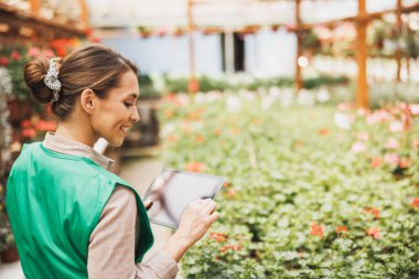 Beautiful young woman working at a greenhouse and writing on a tablet clipboard.