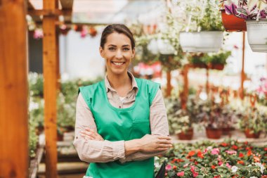 Young woman entrepreneur standing in front of tables of potted flowers and smiling at the camera. She is wearing a green apron and working in plant nursery. 