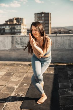 Young woman practicing yoga on a rooftop terrace. City buildings in the background.