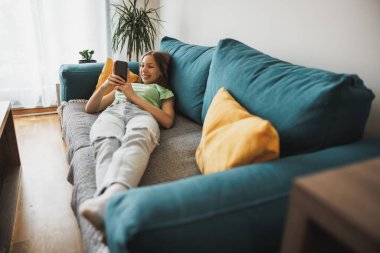 Beautiful teenager girl surfing social media on smart phone while having leisure time at her home.