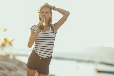 Cheerful teen girl having fun and drinking non alcoholic beer on the beach party by the sea. 