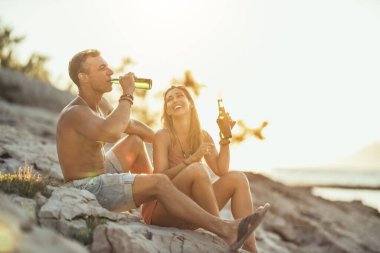 Young couple having fun and drinking beer on the beach by the sea. Enjoying in beautiful sunset and their love.