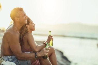Young couple having fun and drinking beer on the beach by the sea. Enjoying their love.