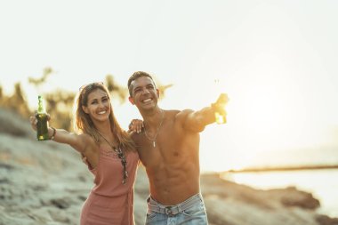 Young couple at the beach having fun, laughing and drinking beer on the beach by the sea. Enjoying in beautiful sunset and their love.