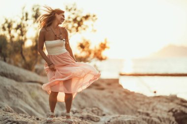 An attractive young woman dressed summer dress is having fun and dancing while enjoying sunset at the beach.