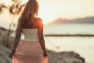 Rear view of an attractive young woman in dress is enjoying summer sunset at the wonderful seaside.